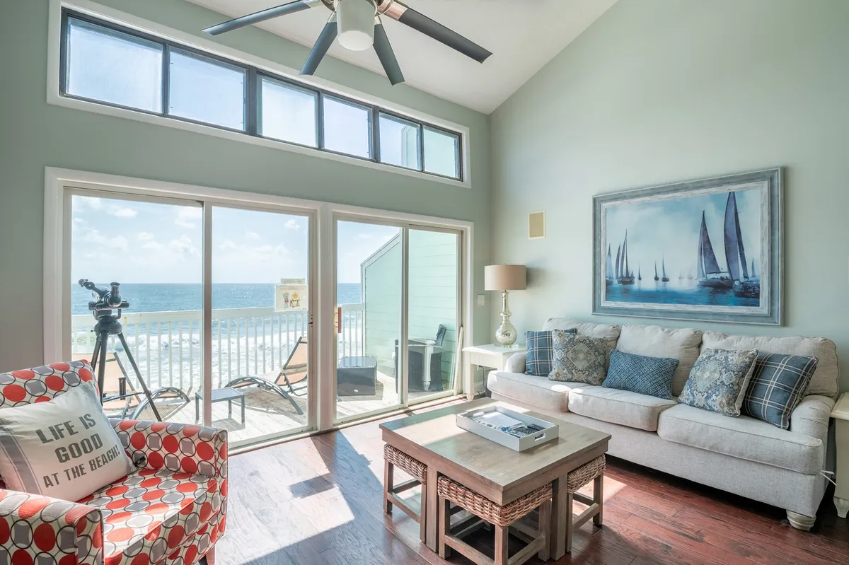 A bright, modern living room with plush seating faces expansive windows that frame a breathtaking ocean view. Sunlight pours in, illuminating coastal-inspired décor, including a colorful accent chair, artistic sailboat prints, and a stylish lamp. The inviting space exudes tranquility, inviting guests to relax and enjoy the scenery from the comfort of the couch or step outside to watch the sunset from the private balcony.