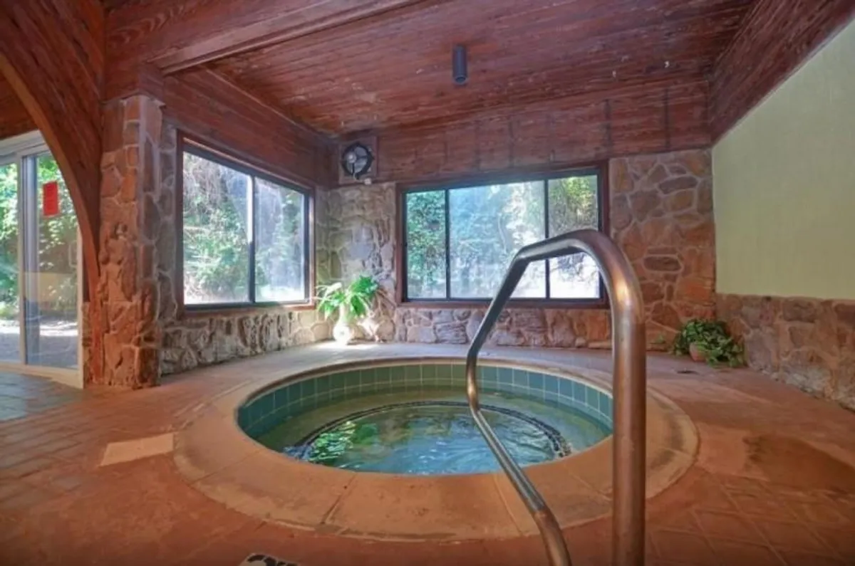 A serene indoor hot tub area featuring a stone wall, large windows that let in natural light, a wooden ceiling, and lush indoor plants. The mood is tranquil and spa-like, perfect for unwinding after a day of adventure.