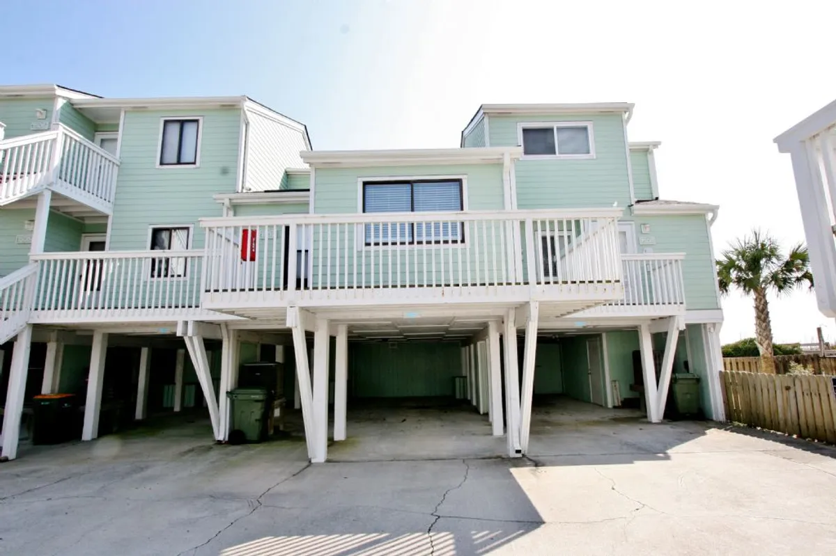 This exterior image displays the building's balcony, palm trees, and the covered area underneath, which is clearly used for parking vehicles. The perspective shows how the assigned covered parking is situated beneath the building, giving guests a sense of entry and convenient access.