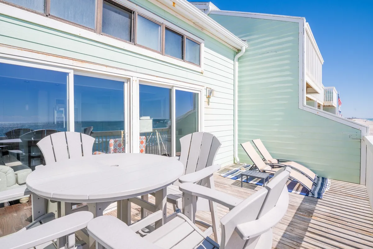 On the private sundeck, an outdoor dining table and lounge chairs are arranged to take full advantage of the large windows and scenic oceanfront view. The connection between indoor and outdoor living is showcased, inviting relaxation and alfresco meals.