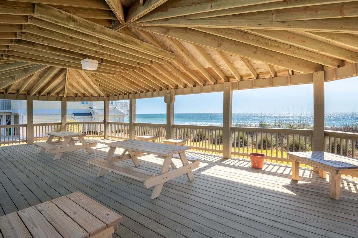 A cluster of wooden picnic tables set on open ground with an expansive ocean view in the background. The scene feels relaxed and social, perfect for gatherings, picnics, or enjoying a meal after a day of exploring local attractions. The wide-open space emphasizes Kure Beach’s coastal charm and opportunities for outdoor activities.