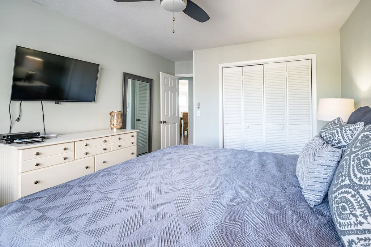 A tranquil bedroom scene featuring a bed with patterned bedding, a television mounted on the wall, a dresser with decorative accents, and a mirror reflecting natural light. The mood is cozy and inviting, ideal for relaxation and private downtime.