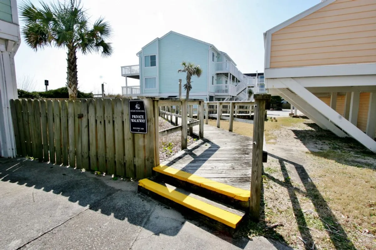 A casual wooden walkway flanked by a palm tree, leading out to the beach. Bright yellow steps and a cheerful fence with a sign invite guests onto the sand. The scene captures the transition from the property to the wide open beach beyond, with sunlight suggesting a warm, welcoming day for outdoor adventure.