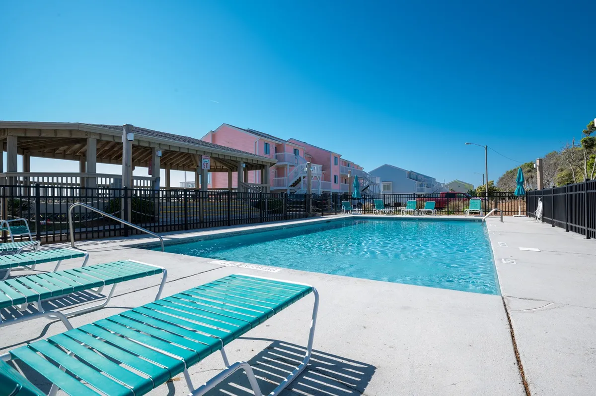 An inviting outdoor swimming pool area bordered by pink resort buildings and a covered section for shade. Lounge chairs line the perimeter, providing a relaxing spot for sunbathers and social gatherings, while the pool itself offers ample space for swimming and play.