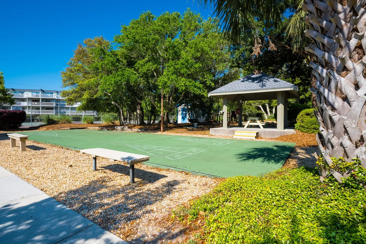 A vibrant outdoor activity space featuring a green playing surface, a quaint gazebo, benches, and lush trees. The environment is cheerful and inviting, suggesting opportunities for games, picnics, and social events—all against the backdrop of leafy island greenery.