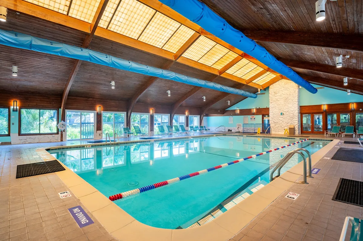A serene indoor swimming pool with a wooden ceiling, large windows letting in natural light, and comfortable poolside lounge chairs. The scene exudes tranquility and is ideal for year-round swimming and relaxation.